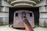 A large, rusted artillery gun protrudes from a fortified concrete bunker structure. The setting has a historical and abandoned feel, with the gun showing signs of wear and age. The bunker is constructed with heavy concrete and the gun extends outward, suggesting it was once used for defensive purposes.