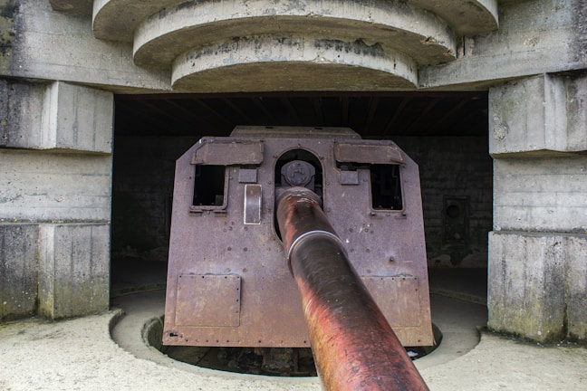 A large, rusted artillery gun protrudes from a fortified concrete bunker structure. The setting has a historical and abandoned feel, with the gun showing signs of wear and age. The bunker is constructed with heavy concrete and the gun extends outward, suggesting it was once used for defensive purposes.