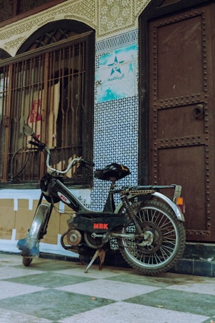 A vintage MBK motorbike leans against a wall adorned with intricate tile patterns and an arched wooden door. The tiles display a mix of geometric designs in blue and white, with a worn sign featuring Arabic script. The motorbike has a metal frame, visible engine parts, and a chain, indicating a well-used condition. The scene gives a sense of an urban, perhaps historical, setting.