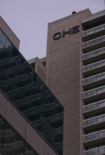 A tall building with a concrete facade featuring the letters 'CHE' near the top. The structure to the right has balconies with white railings, and the building on the left has a glass exterior with reflections.