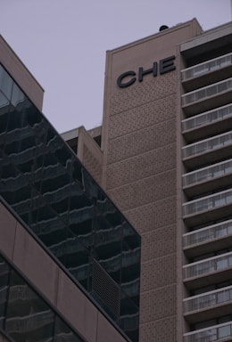 A tall building with a concrete facade featuring the letters 'CHE' near the top. The structure to the right has balconies with white railings, and the building on the left has a glass exterior with reflections.