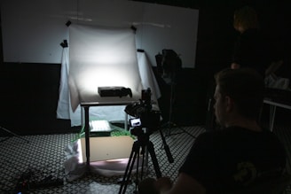 A dimly lit photography setup with a lightbox on a table, aimed by a camera mounted on a tripod. The background is dark with another person standing partially visible. The room has a patterned floor and various photography equipment scattered around.