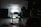 A dimly lit photography setup with a lightbox on a table, aimed by a camera mounted on a tripod. The background is dark with another person standing partially visible. The room has a patterned floor and various photography equipment scattered around.