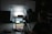 A dimly lit photography setup with a lightbox on a table, aimed by a camera mounted on a tripod. The background is dark with another person standing partially visible. The room has a patterned floor and various photography equipment scattered around.