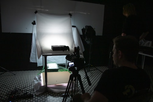 A dimly lit photography setup with a lightbox on a table, aimed by a camera mounted on a tripod. The background is dark with another person standing partially visible. The room has a patterned floor and various photography equipment scattered around.