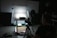 A dimly lit photography setup with a lightbox on a table, aimed by a camera mounted on a tripod. The background is dark with another person standing partially visible. The room has a patterned floor and various photography equipment scattered around.