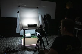 A dimly lit photography setup with a lightbox on a table, aimed by a camera mounted on a tripod. The background is dark with another person standing partially visible. The room has a patterned floor and various photography equipment scattered around.