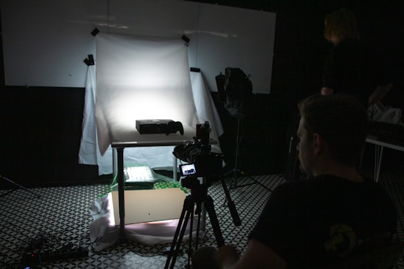 A dimly lit photography setup with a lightbox on a table, aimed by a camera mounted on a tripod. The background is dark with another person standing partially visible. The room has a patterned floor and various photography equipment scattered around.