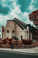 An architectural building with a modern design, featuring circular and arched windows. The structure is surrounded by a brick wall and lush, reddish-brown foliage. The sky is a dramatic shade of blue with scattered clouds, adding to the overall atmosphere.