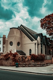 An architectural building with a modern design, featuring circular and arched windows. The structure is surrounded by a brick wall and lush, reddish-brown foliage. The sky is a dramatic shade of blue with scattered clouds, adding to the overall atmosphere.