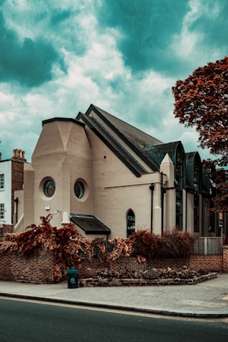 An architectural building with a modern design, featuring circular and arched windows. The structure is surrounded by a brick wall and lush, reddish-brown foliage. The sky is a dramatic shade of blue with scattered clouds, adding to the overall atmosphere.