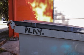 A gray metal dumpster with a black lid has the word 'PLAY.' spray-painted in black on the side. The background features an orange and yellow wall with shadows, and part of a tree is visible to the left.