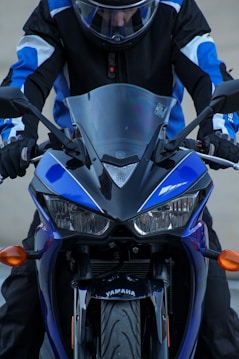 A person wearing a black and blue motorcycle suit and helmet is seated on a Yamaha motorcycle. The focus is on the front view of the motorcycle, highlighting its headlights, handlebars, and the Yamaha logo. The motorcycle exhibits a sleek design with a prominent windshield.