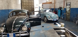 Two vintage Volkswagen Beetles are parked inside a garage with blue and white walls. The garage door is open, allowing natural light to illuminate the space. One car is black with its door open, and the other is light blue. Various items are scattered on the floor, including a pair of shoes, a bucket, and some tools. Bicycles are leaning against the wall on the right side.