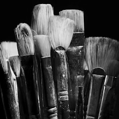A collection of artist paintbrushes with varying bristle sizes and shapes is tightly grouped together. The brushes have worn handles and are set against a dark background, creating a dramatic contrast. The texture of the bristles is highlighted by the monochrome lighting.