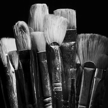 A collection of artist paintbrushes with varying bristle sizes and shapes is tightly grouped together. The brushes have worn handles and are set against a dark background, creating a dramatic contrast. The texture of the bristles is highlighted by the monochrome lighting.