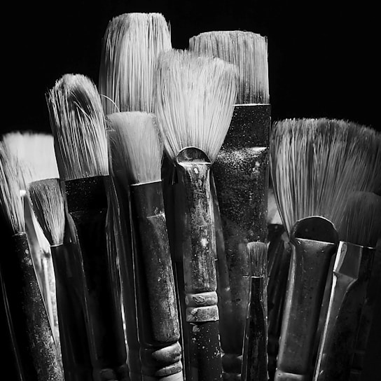 A collection of artist paintbrushes with varying bristle sizes and shapes is tightly grouped together. The brushes have worn handles and are set against a dark background, creating a dramatic contrast. The texture of the bristles is highlighted by the monochrome lighting.