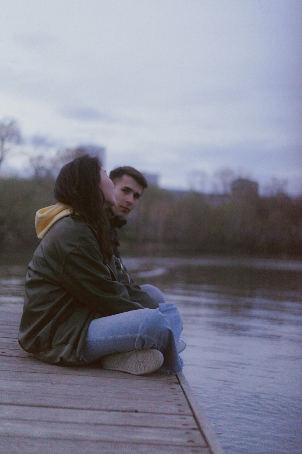 a man and woman sitting on a dock by a lake