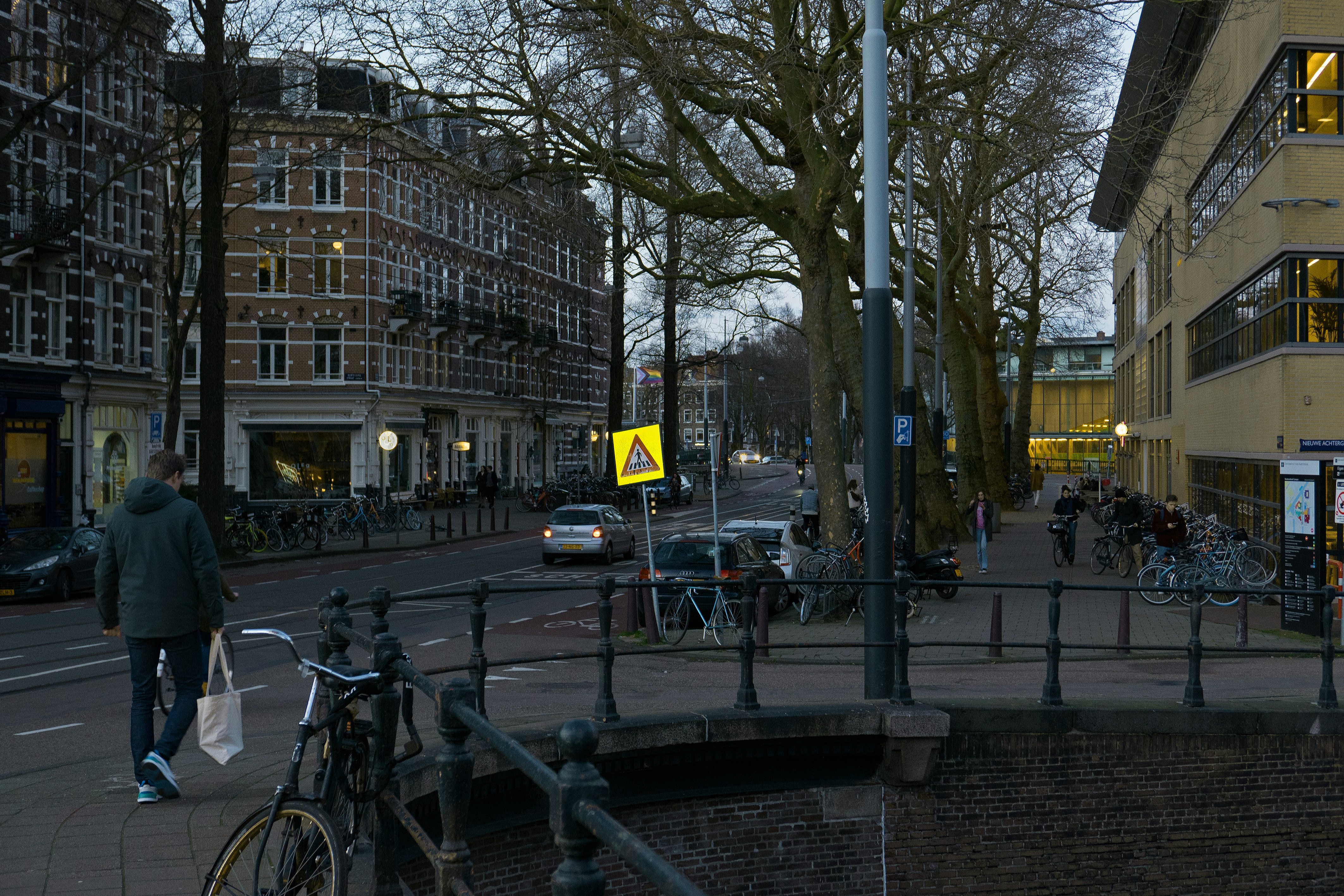 A street view photo with walking people over the Roetersstraat in Amsterdam city with its many bridges over the crossing canals on a gray day. At the left a university campus building called Roeterseiland. It is early evening twilight half February 2022. Free Amsterdam street photography by Fons Heijnsbroek, Netherlands. // Rechtenvrije, gratis foto in hoge resolutie van een student die over de brug loopt bij de Nieuwe Achtergracht,op de Roetersstraat met zijn grote bomen (platanen) - dicht bij de universiteits-campus Roeterseiland in Amsterdam (rechts!); straatfotografie Nederland, 2022.