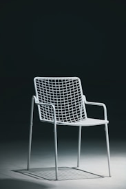 A single white metal chair with a minimalist wireframe design is placed on a smooth, solid surface with a dark background. The lighting creates shadows underneath the chair, emphasizing its geometric structure.