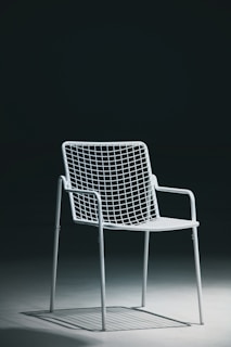 A single white metal chair with a minimalist wireframe design is placed on a smooth, solid surface with a dark background. The lighting creates shadows underneath the chair, emphasizing its geometric structure.