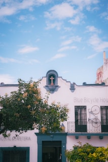 A building labeled 'Blue Bicycle House' with a teal-colored facade features a prominent cross above an arched doorway and a decorative bicycle mounted above the second-floor window. The building has wrought iron railings, and in front of it, there is a leafy tree, all under a clear blue sky with a few clouds.