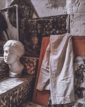 A plaster bust of a woman's head is placed on a mosaic step next to a draped white shirt. The background features textured, weathered walls and a dark abstract painting partially visible. The overall setting is an artist's studio with a rustic and aged ambiance.
