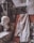 A plaster bust of a woman's head is placed on a mosaic step next to a draped white shirt. The background features textured, weathered walls and a dark abstract painting partially visible. The overall setting is an artist's studio with a rustic and aged ambiance.