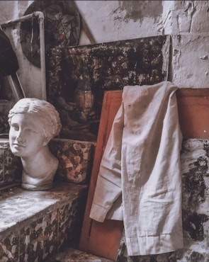 A plaster bust of a woman's head is placed on a mosaic step next to a draped white shirt. The background features textured, weathered walls and a dark abstract painting partially visible. The overall setting is an artist's studio with a rustic and aged ambiance.