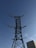 A tall, metal electricity pylon stretches upwards against a clear blue sky. Its intricate lattice structure supports high-voltage power lines that extend in various directions. In the background, a portion of a building is visible in the lower right corner.