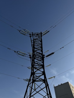 A tall, metal electricity pylon stretches upwards against a clear blue sky. Its intricate lattice structure supports high-voltage power lines that extend in various directions. In the background, a portion of a building is visible in the lower right corner.