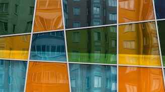 Reflections of a building with multiple windows are visible in a series of colorful, tilted glass panels. The panels are arranged in a grid pattern and display a mix of green, orange, blue, and yellow hues that give a distorted view of the building's architecture.