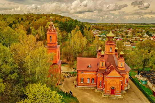 Aerial view of a red-brick Orthodox church with golden domes surrounded by lush greenery and a small village in the background. The architecture is distinct with intricate details, set against a partly cloudy sky.