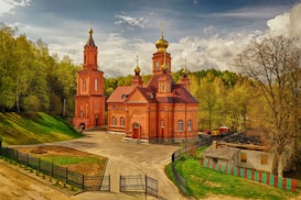 A red brick Orthodox church is set amidst a lush green landscape with a cloudy sky overhead. The architecture features classic domes with golden crosses, and a tall bell tower stands prominently. The surrounding area includes a rustic building, trees, and a paved pathway leading to the entrance.