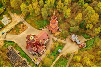 An aerial view of a complex of traditional Russian-style buildings with golden domes surrounded by lush green trees. The central building is a large church with a distinctive roof and multiple smaller domed structures around it. Pathways connect the buildings, and a road is visible with a couple of parked cars.