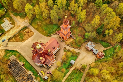 An aerial view of a complex of traditional Russian-style buildings with golden domes surrounded by lush green trees. The central building is a large church with a distinctive roof and multiple smaller domed structures around it. Pathways connect the buildings, and a road is visible with a couple of parked cars.