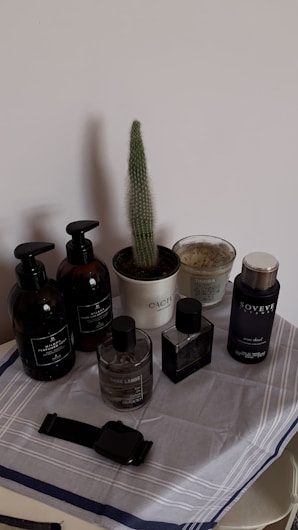 A selection of grooming products arranged on a cloth-covered surface. Included are bottles of various sizes and colors, a wristwatch, and a small potted cactus. The bottles feature a minimalist design, mostly in black and dark hues, and are accompanied by a scented candle.
