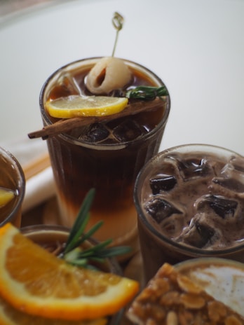 A collection of cold beverages garnished with fruit slices and herbs is arranged on a table. One drink features a lychee on a skewer, a cinnamon stick, and a slice of lemon, while another appears chocolaty with visible ice cubes. The front shows an orange slice, and there's a glimpse of a nutty snack.