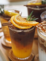A collection of iced beverages with slices of orange and lemon garnishes. Visible cinnamon sticks and greenery add a touch of freshness to the presentation. The drinks appear to be arranged on a wooden surface.