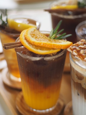 A collection of iced beverages with slices of orange and lemon garnishes. Visible cinnamon sticks and greenery add a touch of freshness to the presentation. The drinks appear to be arranged on a wooden surface.
