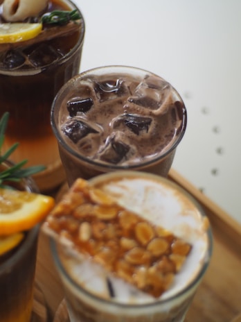 A close-up of three beverages in glasses, each filled with ice cubes. The drink in the foreground has a topping of sliced almonds and caramel. The drink to the left features a slice of citrus and a sprig of rosemary. Each drink displays distinct layers, with the colors transitioning from a creamy top to darker hues at the bottom.