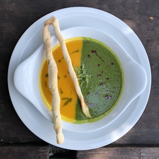 A white bowl containing two different soups, one with a vibrant orange color and the other a rich green. Each soup is garnished with a few drops of oil and fresh herbs. Two long, twisted breadsticks are placed on top, crossing over the bowl. The dish is set on a dark wooden table.