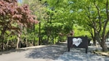 A peaceful road lined with vibrant green trees, accompanied by a sign featuring a heart and the words 'GOOD BYE.' The scene is filled with greenery and the sunlight filters through the leaves, creating a serene atmosphere.