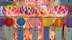A collection of lotus-shaped lanterns in vibrant pink, purple, and yellow hues are arranged above a row of round paper lanterns in various colors. These are displayed with banners featuring Korean calligraphy hanging from each lantern.