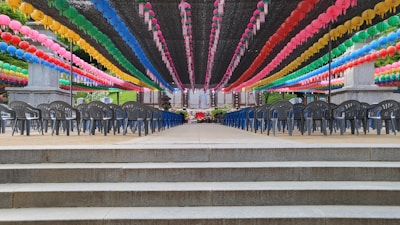 A series of colorful lanterns hang in parallel lines under a canopy, creating a vibrant and festive atmosphere. Rows of empty black chairs are set up in neat lines, suggesting the preparation for an event or ceremony. The setting appears outdoors with steps leading up to the area, and greenery visible in the background.