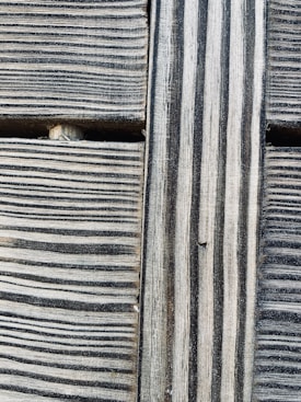 Close-up view of wooden planks with a prominent grain pattern. The wood features alternating light and dark stripes running vertically and horizontally, creating a textured surface. The aged appearance suggests weathering and natural wear, adding a sense of rusticity.