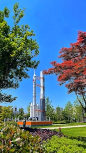 A park scene featuring a large rocket model standing prominently amidst lush greenery and well-maintained landscaping. The rocket is surrounded by trees with vibrant green and red leaves, set against a clear blue sky. In the background, a few buildings peek through the foliage.