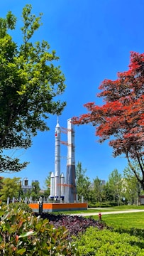 A park scene featuring a large rocket model standing prominently amidst lush greenery and well-maintained landscaping. The rocket is surrounded by trees with vibrant green and red leaves, set against a clear blue sky. In the background, a few buildings peek through the foliage.