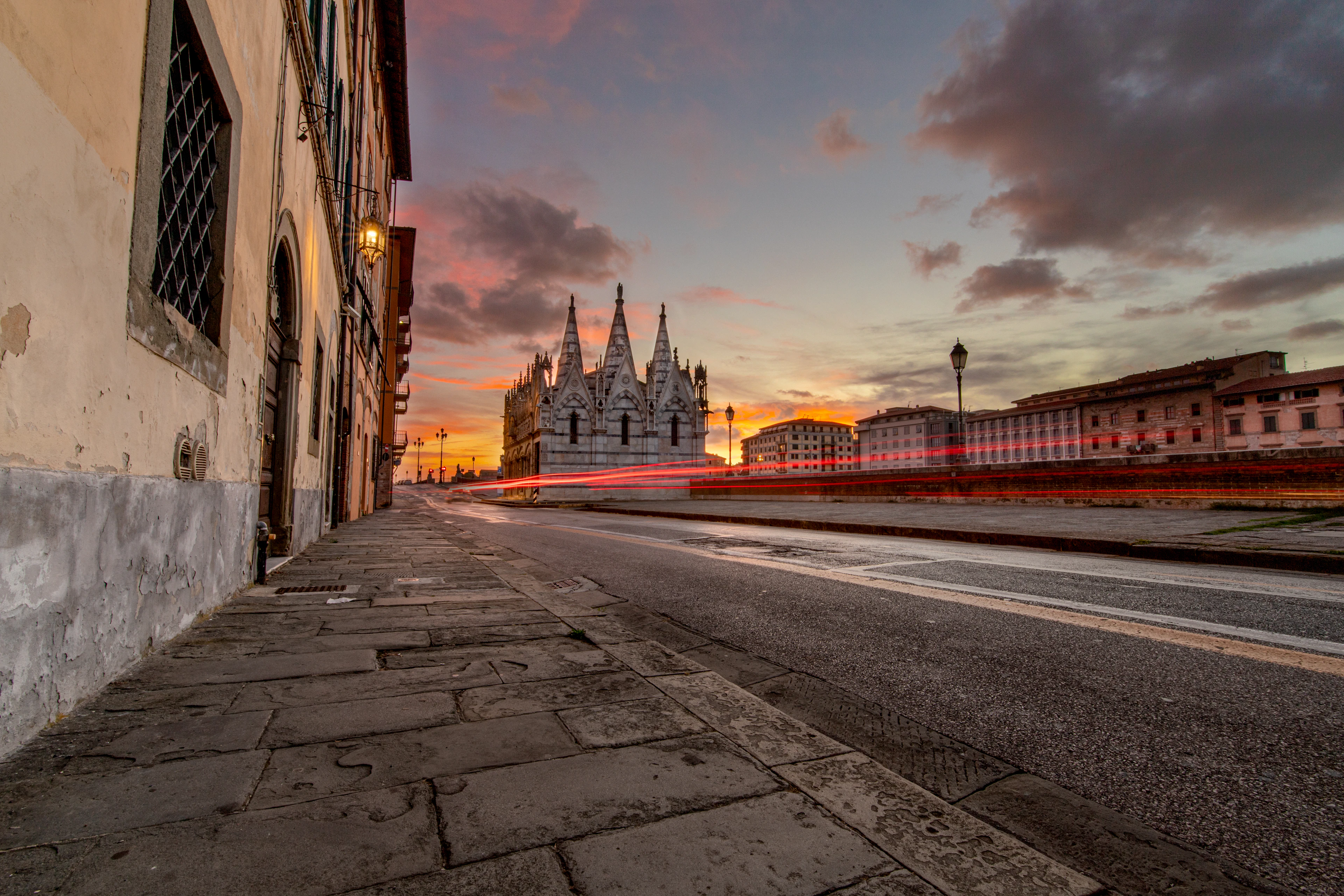 The pinnacled white marble church of Santa Maria della Spina on the Lungarno in Pisa at twilight, its spires silhouetted against a burning orange and violet sky, with lit street lamps and the red streaks of passing traffic along the empty riverside road.