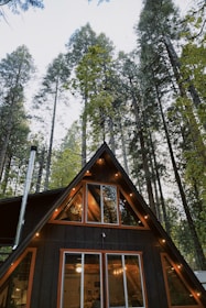 Finished A-frame cabin glowing warmly inside, surrounded by pine trees and a wooden deck.